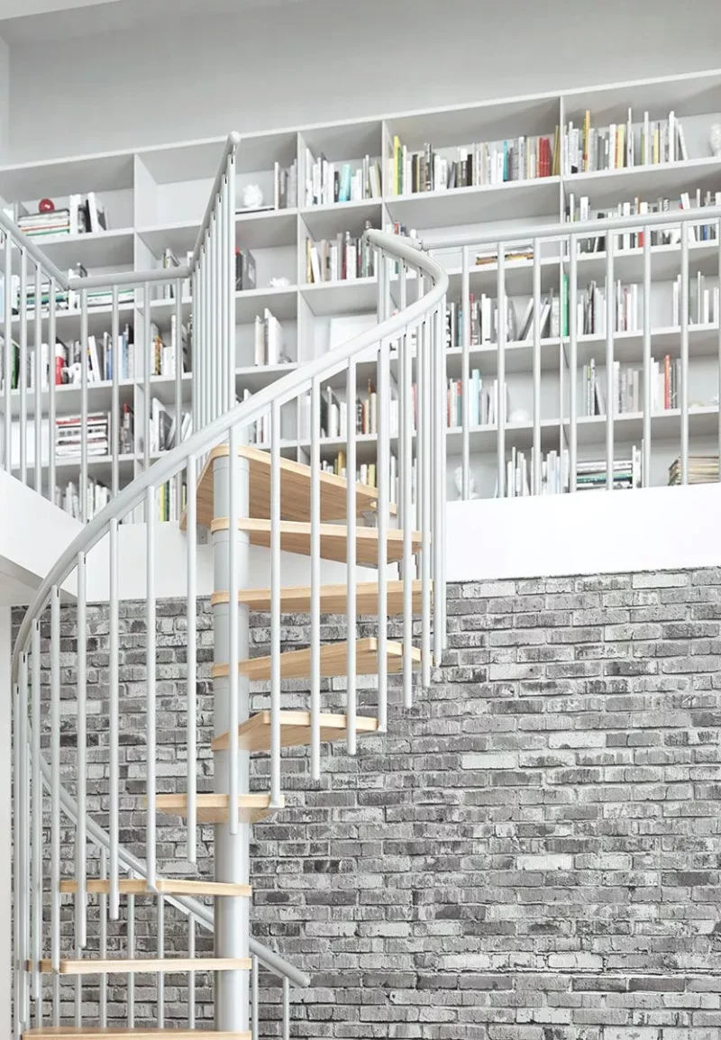 spiralno stubište s metalnim rukohvatom u sivoj boji i drvenim gazištima postavljeno uz zid od sive cigle i bijelu policu s knjigama