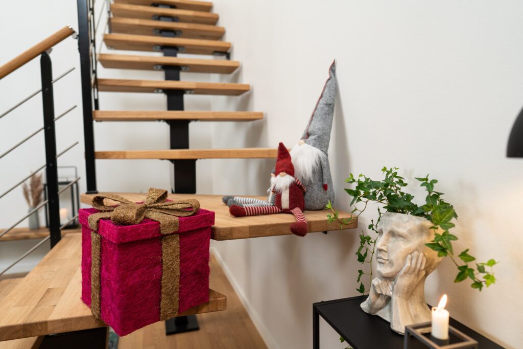 Božićni detalji uz stubište HAMBURG drvene stepenice HAMBURG s crnom čeličnom konstrukcijom, božićni ukrasi poput crvenog poklona i figurica vilenjaka na stubištu, bijeli zid i saksija u obliku lica s biljkom